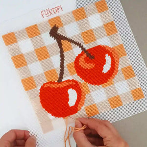 Main réalisant le kit de canevas moderne motif cerises sur fond vichy coloris abricot parfait pour débutants en broderie